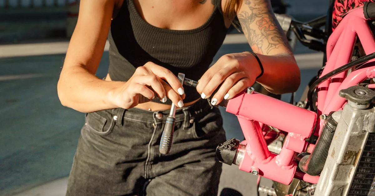 Femme à genoux en train de réparer une moto rose avec des outils en plein air.