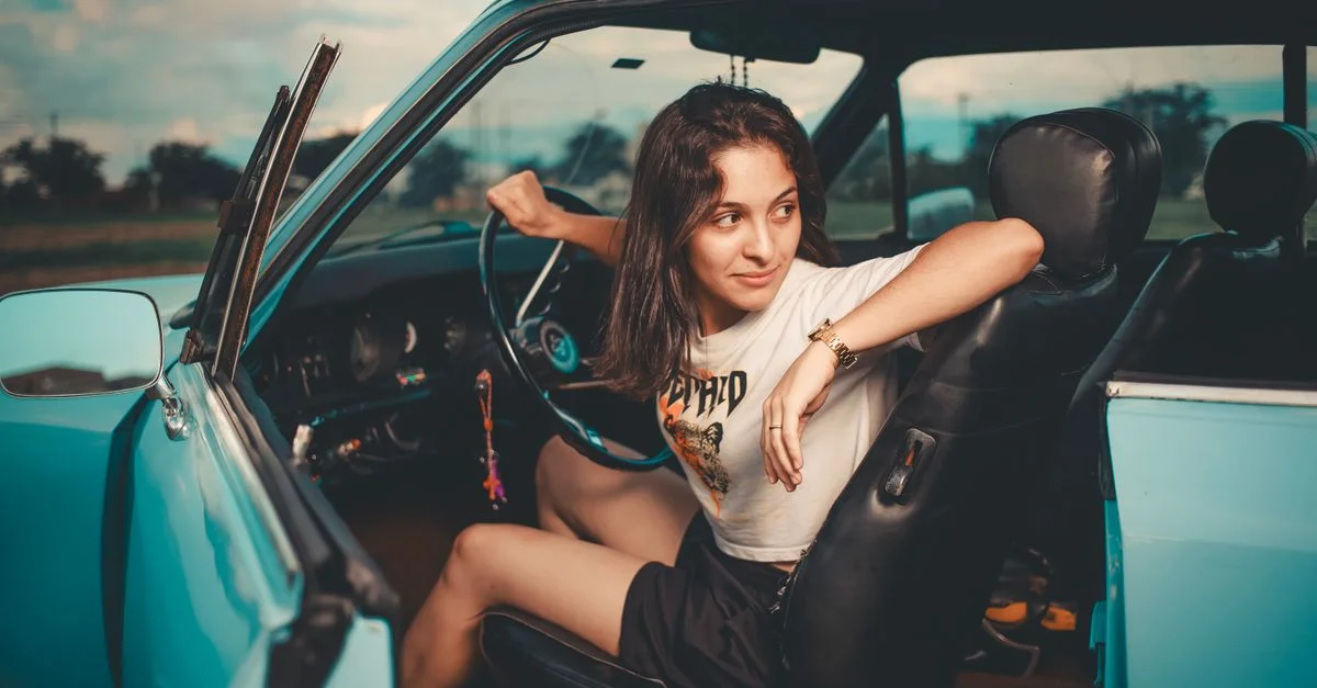 Une jeune femme assise à l'intérieur d'une voiture bleue vintage, profitant du coucher de soleil à l'extérieur.